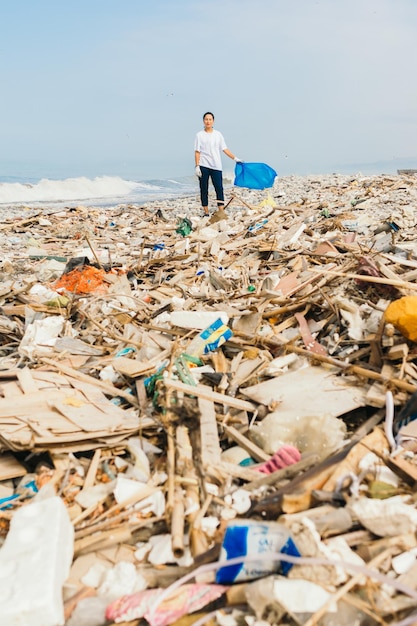 Imagem vertical de uma mulher latina segurando um saco azul sobre uma pilha de lixo e lixo em uma praia poluída. Conceito de poluição ambiental