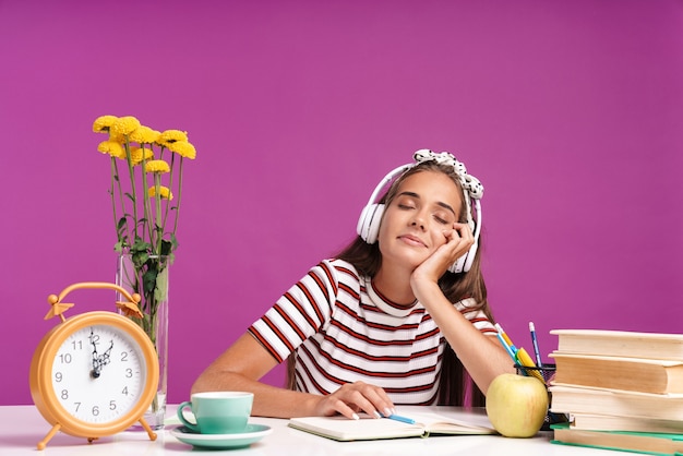 Imagen de agradable chica relajada escuchando música con auriculares mientras está sentado en un escritorio aislado sobre la pared violeta