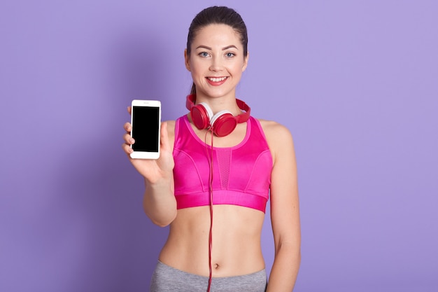 Imagen de agradable sonriente mujer joven linda, sosteniendo teléfono inteligente, con auriculares alrededor del cuello