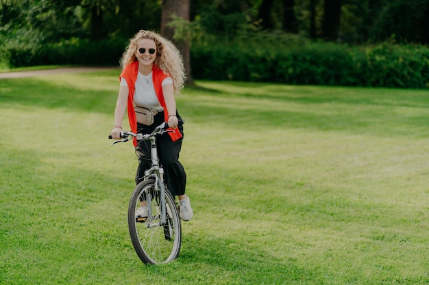 Imagen al aire libre de una joven bastante alegre monta en bicicleta usa gafas de sol ropa informal posa en césped verde pasa tiempo libre en bicicletas de parque en la hermosa naturaleza Concepto de actividad y recreación