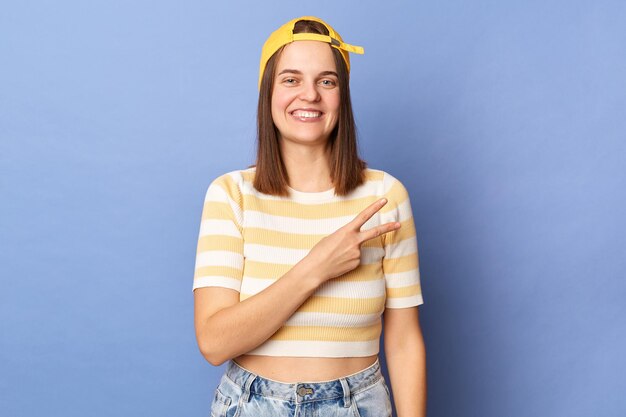 Imagen de una alegre y atractiva adolescente optimista con camiseta a rayas y gorra de béisbol posando aislada sobre fondo azul mostrando el signo de la victoria sonriendo