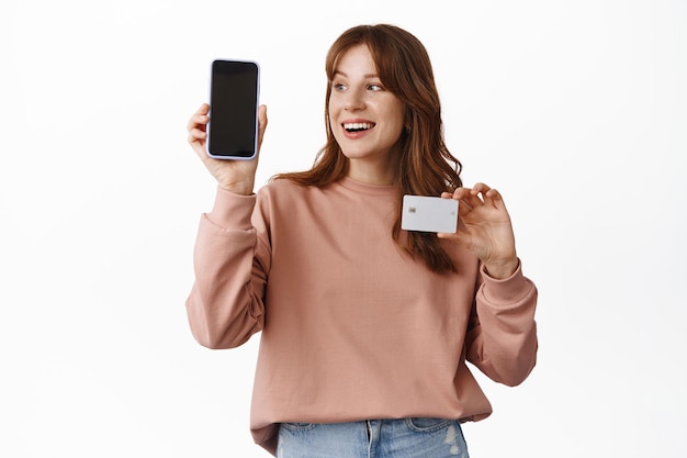 La imagen de una alegre chica pelirroja mira el teléfono inteligente, muestra una pantalla de teléfono vacía y una tarjeta de crédito con una sonrisa complacida, compras en línea, aplicación y concepto de tecnología