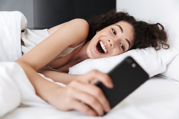 Imagen de alegre mujer caucásica de 20 años con cabello oscuro y rizado acostado en la cama blanca en casa y mirando el teléfono móvil con entusiasmo