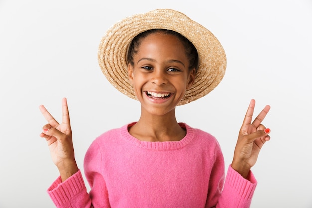 Imagen de la alegre niña afroamericana con sombrero de paja sonriendo y gesticulando el signo de la paz aislado sobre la pared blanca