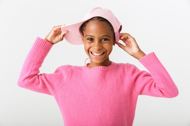 Imagen de una alegre niña afroamericana sonriendo y sosteniendo una gorra rosa aislada sobre una pared blanca