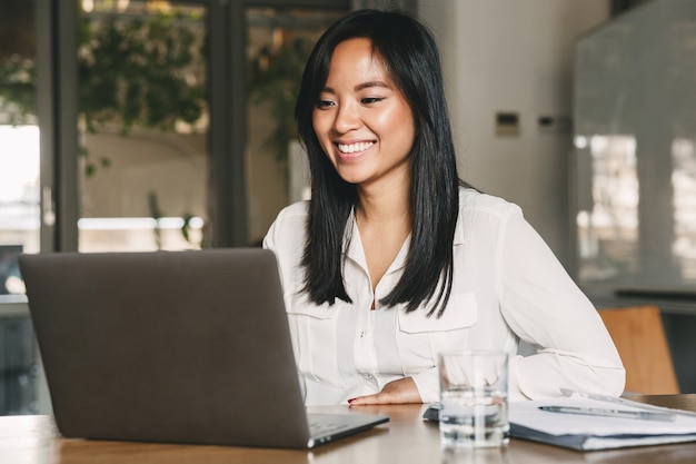 Imagen de alegre trabajadora asiática de 20 años con camisa blanca sonriendo mientras está sentado a la mesa en la oficina y trabajando en la computadora portátil