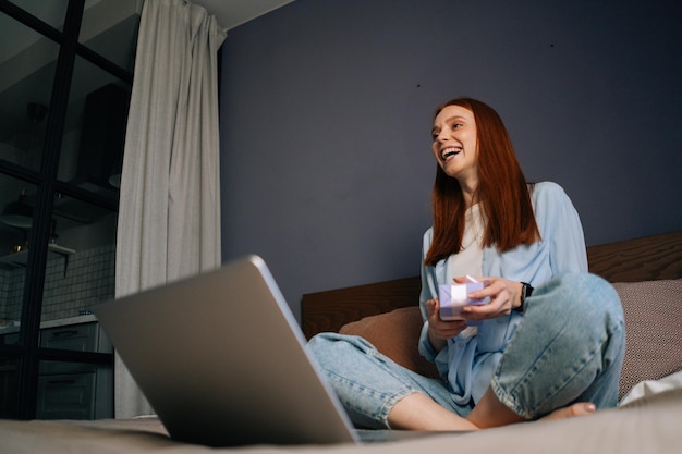 Imagen en ángulo bajo de una joven pelirroja que se ríe con ropa de casa abriendo una caja de regalo con un regalo mientras hace una videollamada en una laptop sentada en la cama en casa en un dormitorio claro.