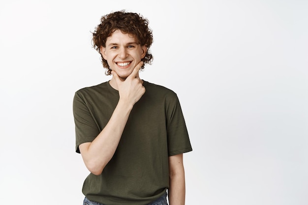 Imagen de un apuesto chico de pelo rizado sonriente tocándose la barbilla y luciendo intrigado pensando en algo parado sobre fondo blanco