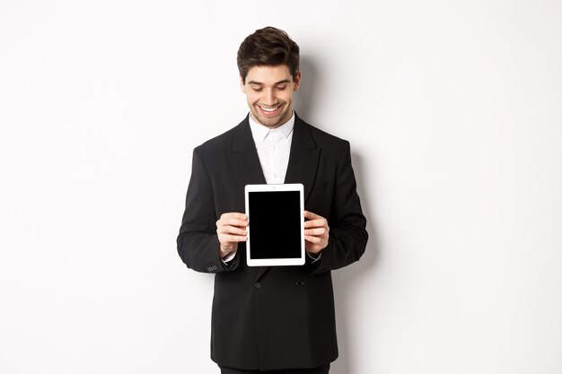 Imagen de un apuesto empresario con traje negro, mirando hacia la pantalla de la tableta digital y mostrando publicidad, de pie contra el fondo blanco.