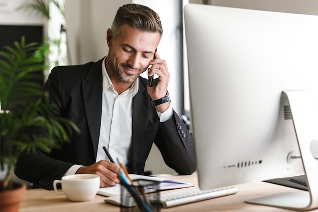 Foto imagen del apuesto hombre de negocios de 30 años con traje hablando por teléfono celular mientras trabaja en la computadora en la oficina