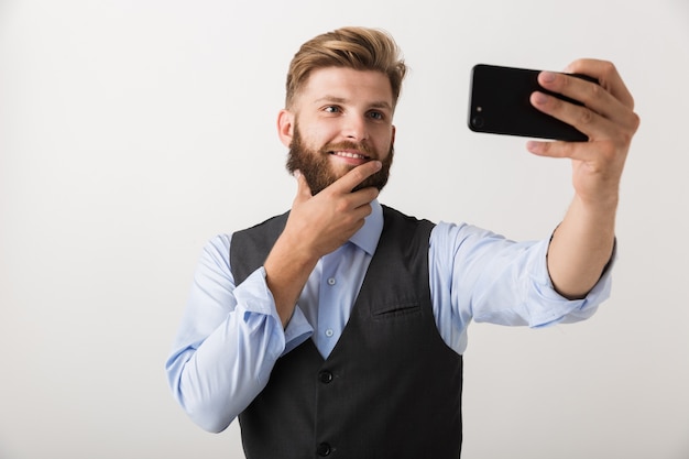 Imagen de un apuesto joven barbudo que se encuentra aislado sobre una pared blanca, tome un selfie por teléfono móvil.