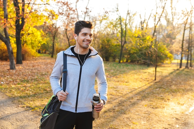 Imagen de un apuesto joven deportista al aire libre en el parque escuchando música con auriculares caminando con bolsa.