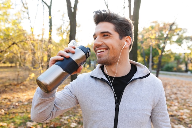 Imagen de un apuesto joven deportista emocional al aire libre en el parque escuchando música con auriculares agua potable.
