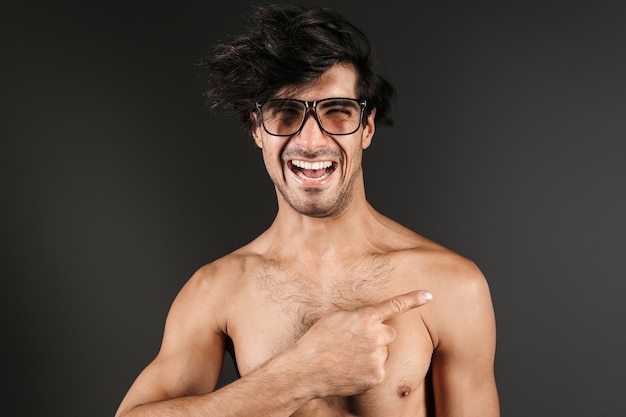 Imagen de un apuesto joven desnudo sonriente posando aislado con gafas apuntando.