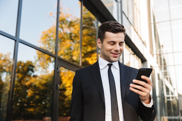 Imagen de un apuesto joven empresario feliz caminando al aire libre cerca del centro de negocios mediante teléfono móvil.