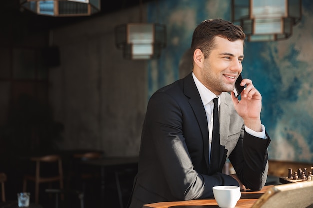 Imagen de un apuesto joven empresario feliz sentado en la cafetería hablando por teléfono móvil.