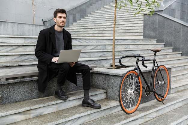 Imagen del apuesto joven empresario sentado al aire libre con ordenador portátil.