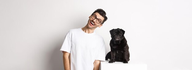Imagen de un apuesto joven parado cerca de un lindo pug negro y un dueño de perro sonriente pasando tiempo con hola