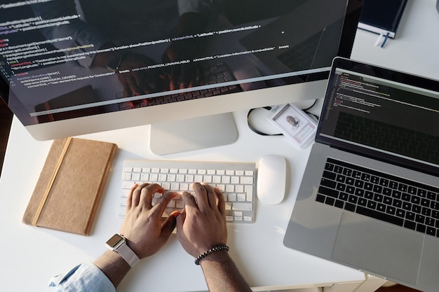 Imagen de arriba hacia abajo de manos masculinas irreconocibles escribiendo en un teclado blanco plateado en la mesa