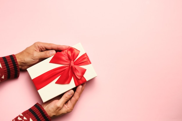 Imagen de arriba de manos sosteniendo un regalo con lazo rojo sobre fondo rosa