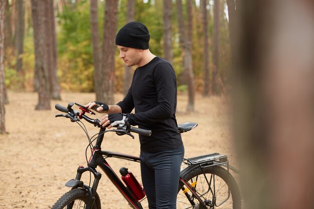 Imagen del atento hombre aventurero de pie en medio del bosque, sosteniendo su bicicleta, mirando la pantalla del teléfono inteligente, buscando el camino de regreso, usando el GPS, eligiendo el camino más corto.