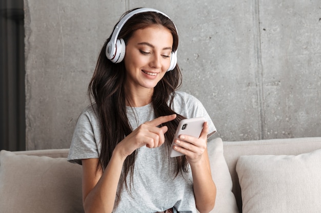 Foto imagen de la atractiva joven morena en auriculares sosteniendo el teléfono inteligente mientras está sentado en el sofá en casa