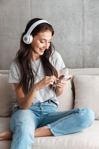 Foto imagen de la atractiva joven morena en auriculares sosteniendo el teléfono inteligente mientras está sentado en el sofá en casa