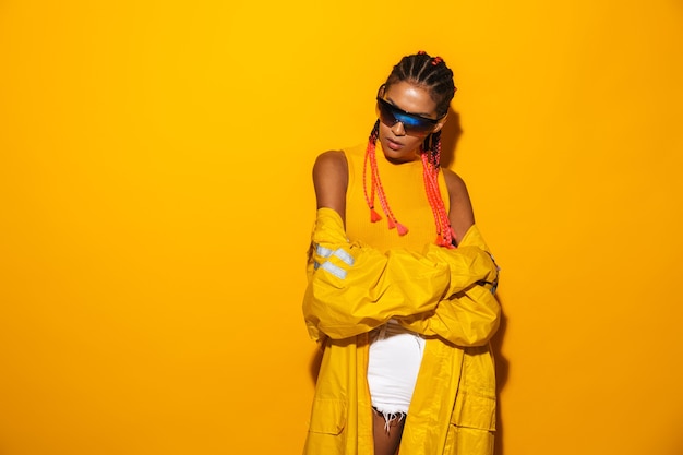 Imagen de atractiva mujer afroamericana con gafas de sol y rompevientos posando aislado sobre pared amarilla