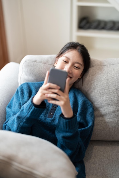 Imagen de una atractiva mujer asiática sonriente usando un teléfono móvil mientras está acostada en un sofá en casa