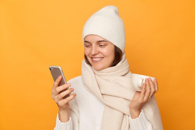 Imagen de una atractiva mujer caucásica alegre con sombrero de suéter y bufanda sosteniendo una taza de café en las manos y usando un teléfono celular leyendo buenas noticias en internet posando aislada sobre un fondo amarillo