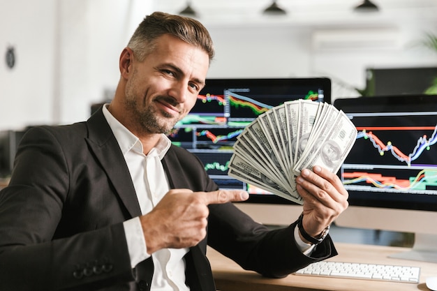 Imagen del atractivo empresario de 30 años vistiendo traje sosteniendo ventilador de dinero mientras trabajaba en la oficina con gráficos y tablas en la computadora