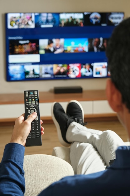 Foto imagen desde atrás de un hombre irreconocible sentado en el sofá con las piernas estiradas buscando contenido para ver en la televisión