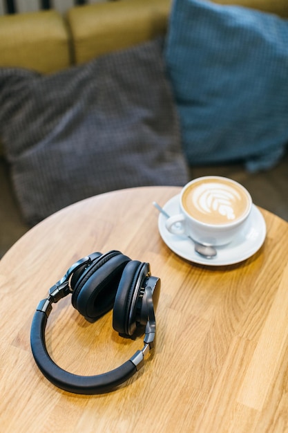 Imagen de auriculares inalámbricos negros y una taza de café blanca en una mesa de madera disfrutando del café