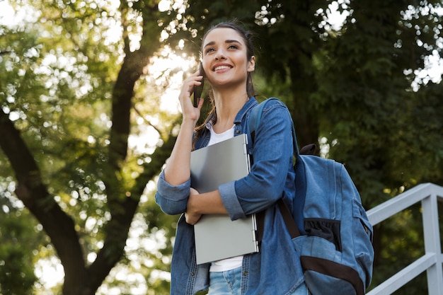 Foto imagen de bastante joven mujer caucásica de pie al aire libre con ordenador portátil hablando por teléfono móvil.