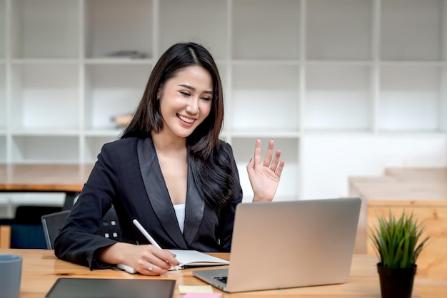 Imagen de una bella empresaria asiática saludando en línea a través de un portátil con lápiz y cuaderno en la oficina.