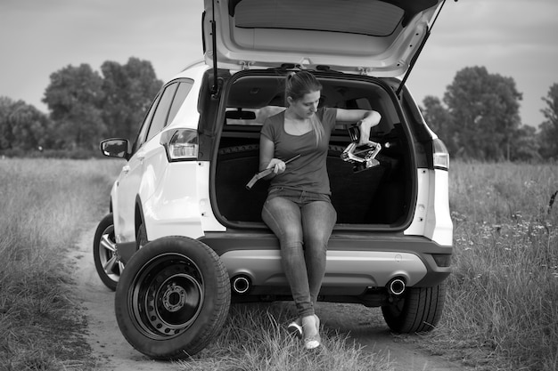 Imagen en blanco y negro de una joven frustrada sentada en un coche roto mirando las herramientas para cambiar las ruedas