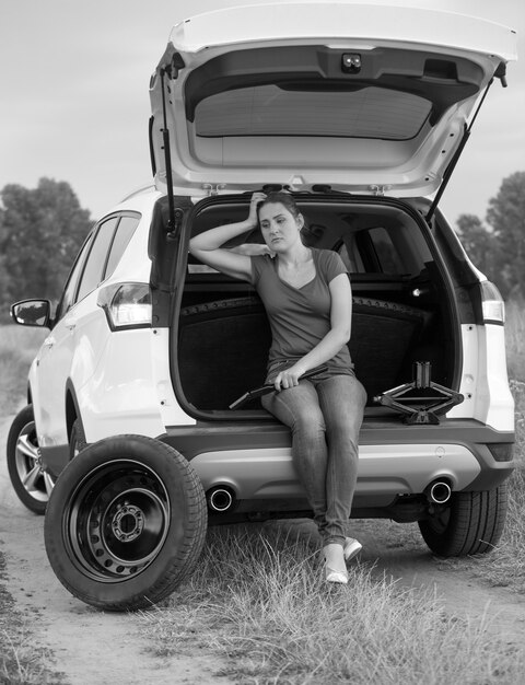 Imagen en blanco y negro de una joven mujer molesta sentada en el maletero del coche abierto y esperando ayuda para cambiar el neumático