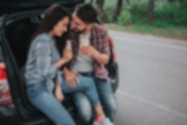 Imagen borrosa de una pareja joven sentada en el maletero y sosteniendo rollos en sus manos. Guy está mirando a la niña. Ella está apoyada en su hombro. Se ven felices