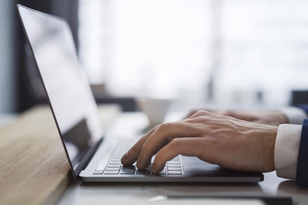 Foto la imagen captura las manos de un hombre de cerca mientras escribe en un elegante teclado de computadora portátil con un entorno de oficina que aparece borroso en el fondo.