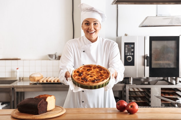Imagen de chef mujer satisfecha con uniforme blanco, posando en la cocina del café con productos horneados