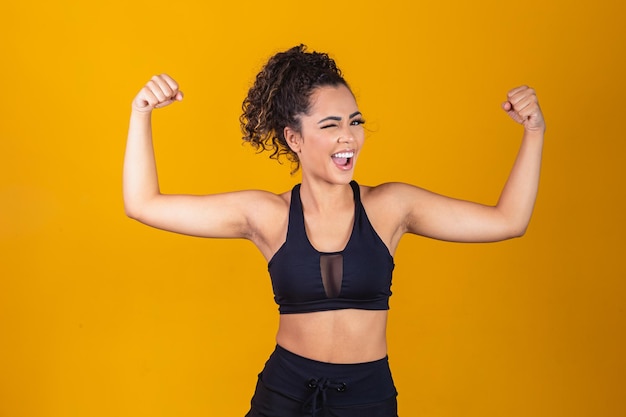 Imagen de una chica afroamericana fuerte y segura, flexionando los bíceps y luciendo segura de sí misma, de pie sobre un fondo amarillo