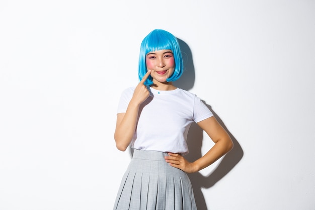 Imagen de una chica asiática kawaii con peluca azul, sonriendo y señalando sus hoyuelos, de pie, vestida para la fiesta de halloween.