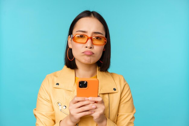 Imagen de una chica asiática triste con gafas de sol que parece decepcionada sosteniendo un teléfono móvil de pie sobre un fondo azul