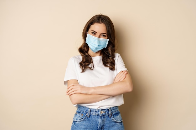 Imagen de una chica enojada con mascarilla médica haciendo muecas y luciendo insultada de pie disgustada contra...