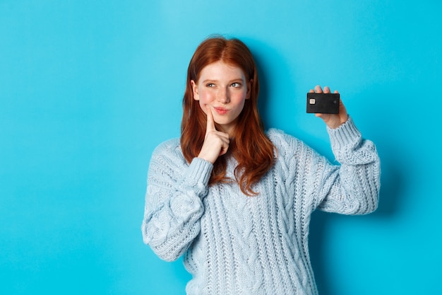 Imagen de una chica pelirroja pensativa pensando en ir de compras, mostrando la tarjeta de crédito y reflexionando, de pie sobre fondo azul.