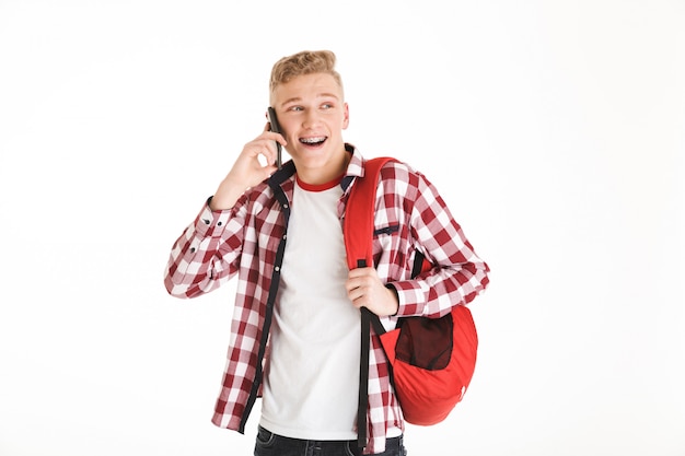 Imagen de chico alegre hipster vistiendo camisa a cuadros y mochila hablando por teléfono móvil con una sonrisa, aislado sobre la pared blanca