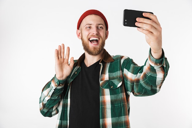 Imagen de chico caucásico con sombrero y camisa a cuadros tomando fotos selfie en teléfono celular, mientras está de pie aislado sobre fondo blanco.