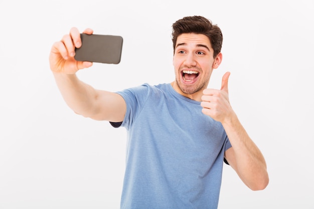 Imagen de chico feliz en estudio sonriendo y mostrando el pulgar hacia arriba mientras se fotografía en un teléfono inteligente negro, aislado sobre la pared blanca