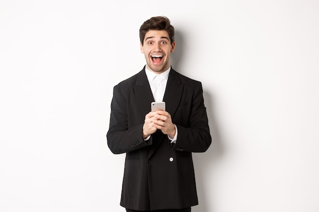 Foto imagen de un chico guapo emocionado recibiendo una oferta promocional en el teléfono móvil, mirando sorprendido y sonriendo, de pie contra el fondo blanco.