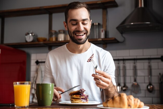 Imagen de chico morena de 30 años sentado en la mesa y comiendo mientras desayuna en un apartamento moderno